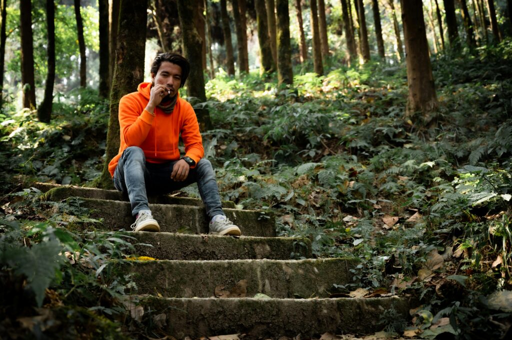 Man in an orange hoodie sits smoking on forest steps in Tukdah, India, surrounded by lush greenery.