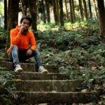 Man in an orange hoodie sits smoking on forest steps in Tukdah, India, surrounded by lush greenery.