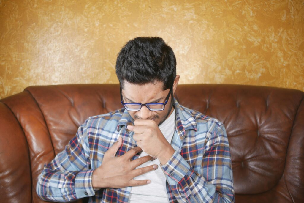 A young man in glasses and plaid shirt coughing, seated on a leather sofa indoors.