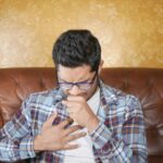 A young man in glasses and plaid shirt coughing, seated on a leather sofa indoors.