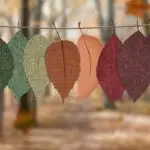 Fabric leaves hang on a rope, showcasing autumn colors in a forest backdrop.