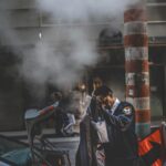 A security guard stands beside a car adjusting his coat on a steamy urban street.