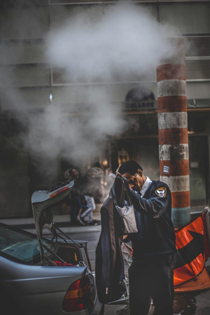 A security guard stands beside a car adjusting his coat on a steamy urban street.