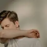 A young man sneezes into his elbow, showcasing a hygiene practice, in an indoor setting.