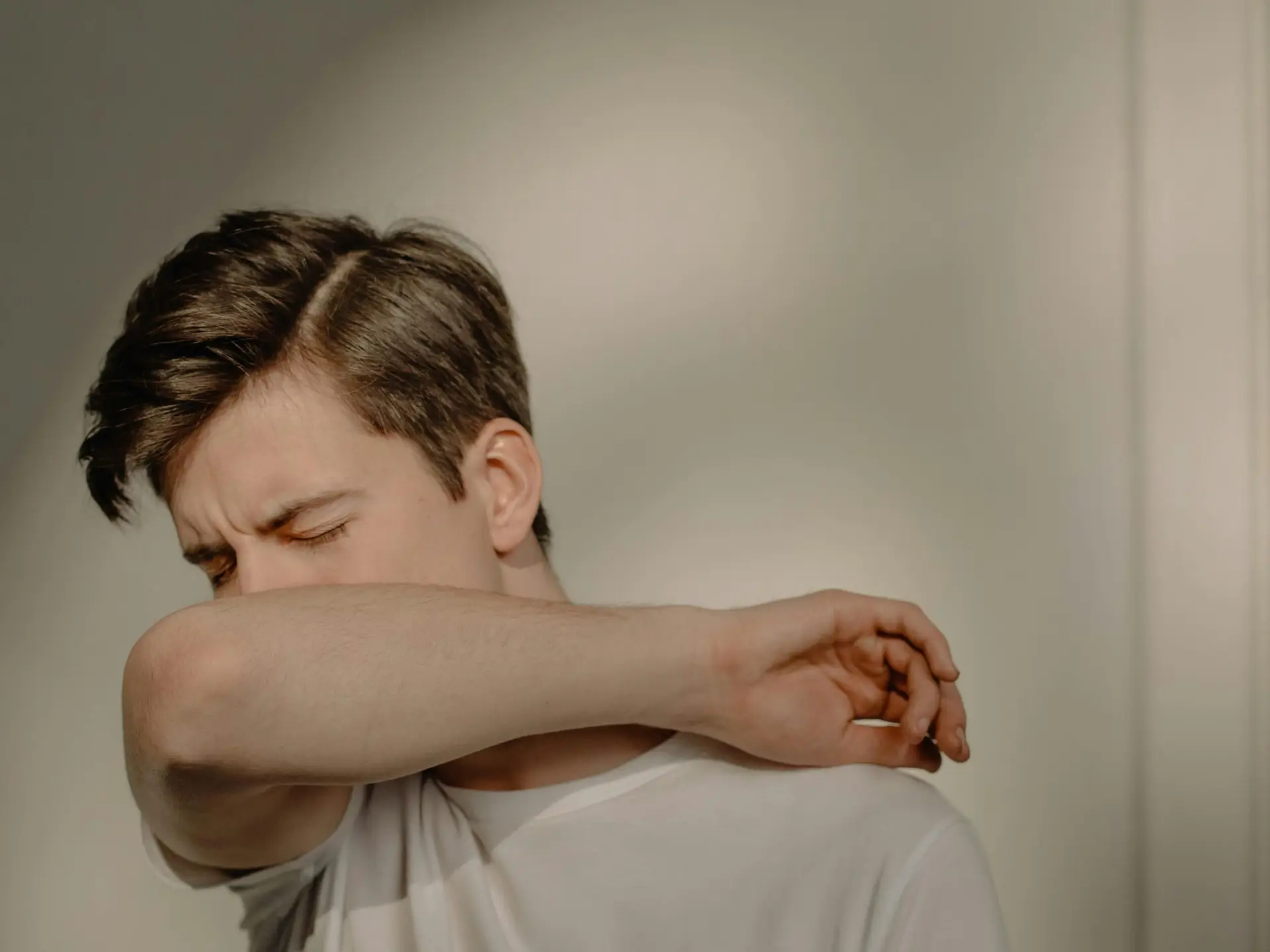 A young man sneezes into his elbow, showcasing a hygiene practice, in an indoor setting.