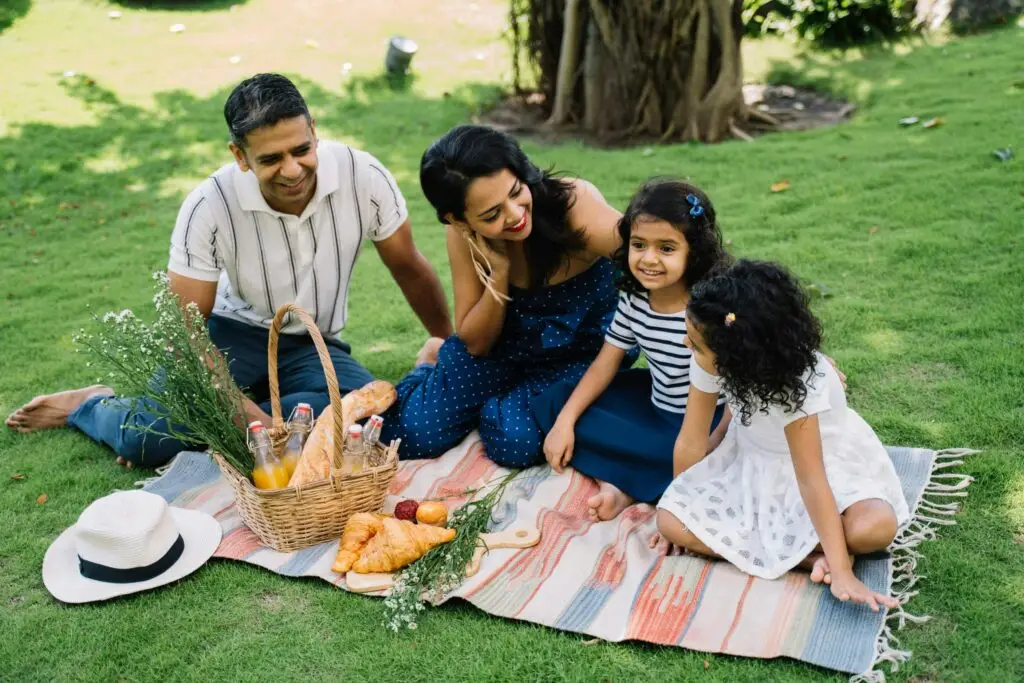 A family of four enjoys a sunny picnic on green grass with smiles and food.