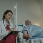 A female doctor checks a medical chart beside a male patient in a hospital room.