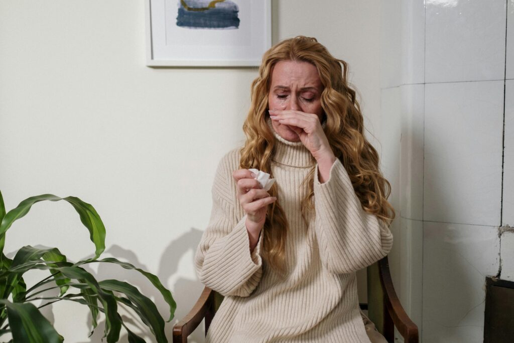 A woman with a runny nose sitting indoors, using a tissue to relieve her allergies.