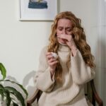 A woman with a runny nose sitting indoors, using a tissue to relieve her allergies.