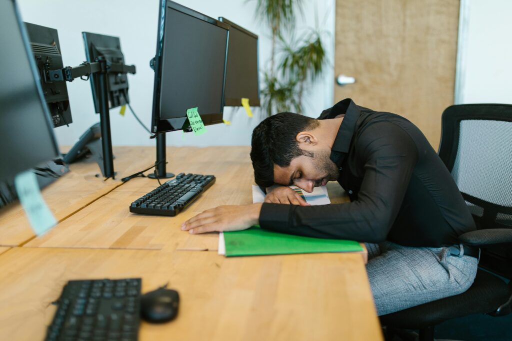 An exhausted office worker resting on a desk surrounded by computer monitors in a modern workspace.