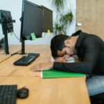 An exhausted office worker resting on a desk surrounded by computer monitors in a modern workspace.