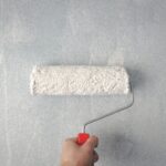 A person's hand using a paint roller to apply white paint on a wall.