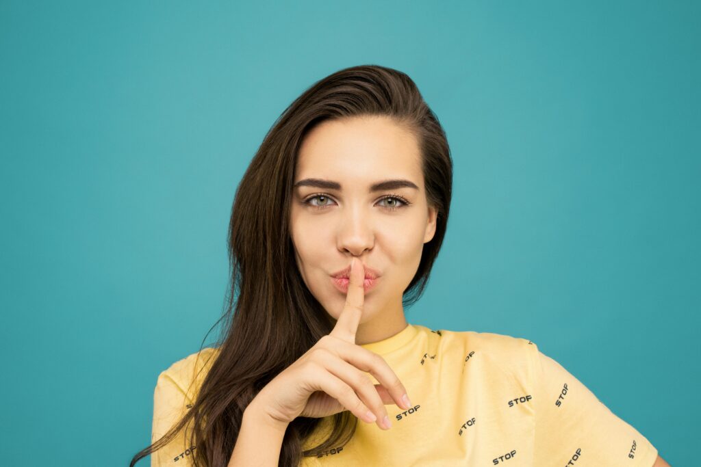 Portrait of a woman with brown hair posing with a hush gesture against a blue background.