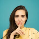 Portrait of a woman with brown hair posing with a hush gesture against a blue background.