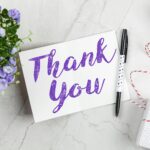A stylish thank you card paired with purple flowers, a gift box, and pen on a marble surface.