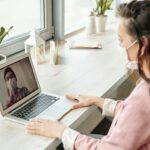 Woman and doctor video calling on laptop, both wearing masks, symbolizing telehealth during COVID-19.