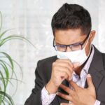 Asian man coughing in a black suit with eyeglasses and face mask indoors.